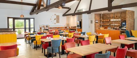 A modern dining area with wooden tables and colorful chairs, a kitchen in the background, and exposed beams on the ceiling.