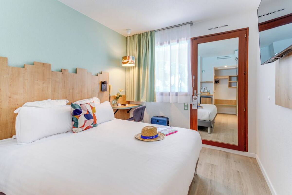 Une chambre d’hôtel avec un lit, un bureau, une chaise, une valise et un miroir qui reflète un coin cuisine.