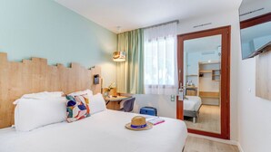 A hotel room with a bed, a desk, a chair, a suitcase, and a mirror reflecting a kitchenette.