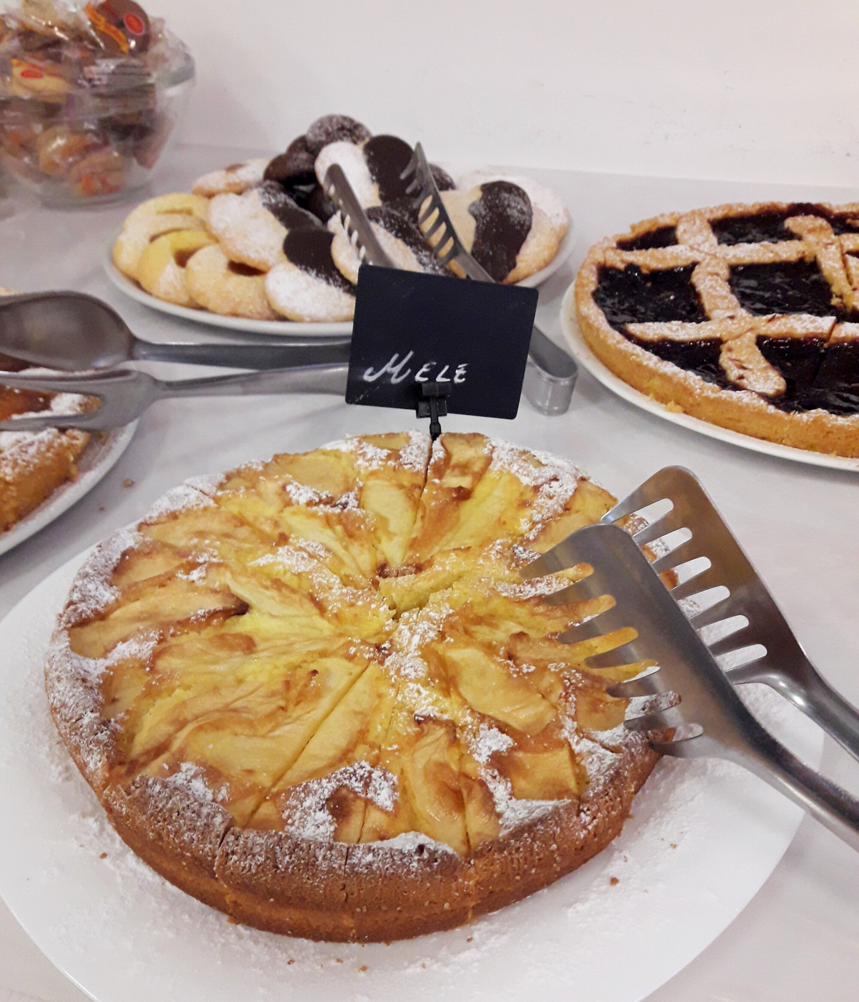 Uma mesa de sobremesas com diversos tortas e doces, incluindo uma torta de maçã em destaque, com uma placa que diz "Mele".
