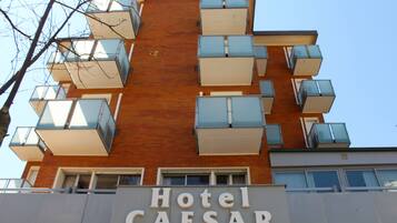 Un hotel llamado Hotel Caesar con un diseño arquitectónico moderno, que cuenta con varios balcones y un cielo despejado al fondo.