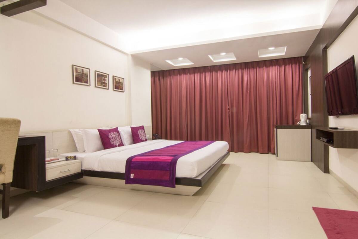 Una habitación de hotel moderna con cama, mesitas de noche, televisor y un gran ventanal con cortinas.