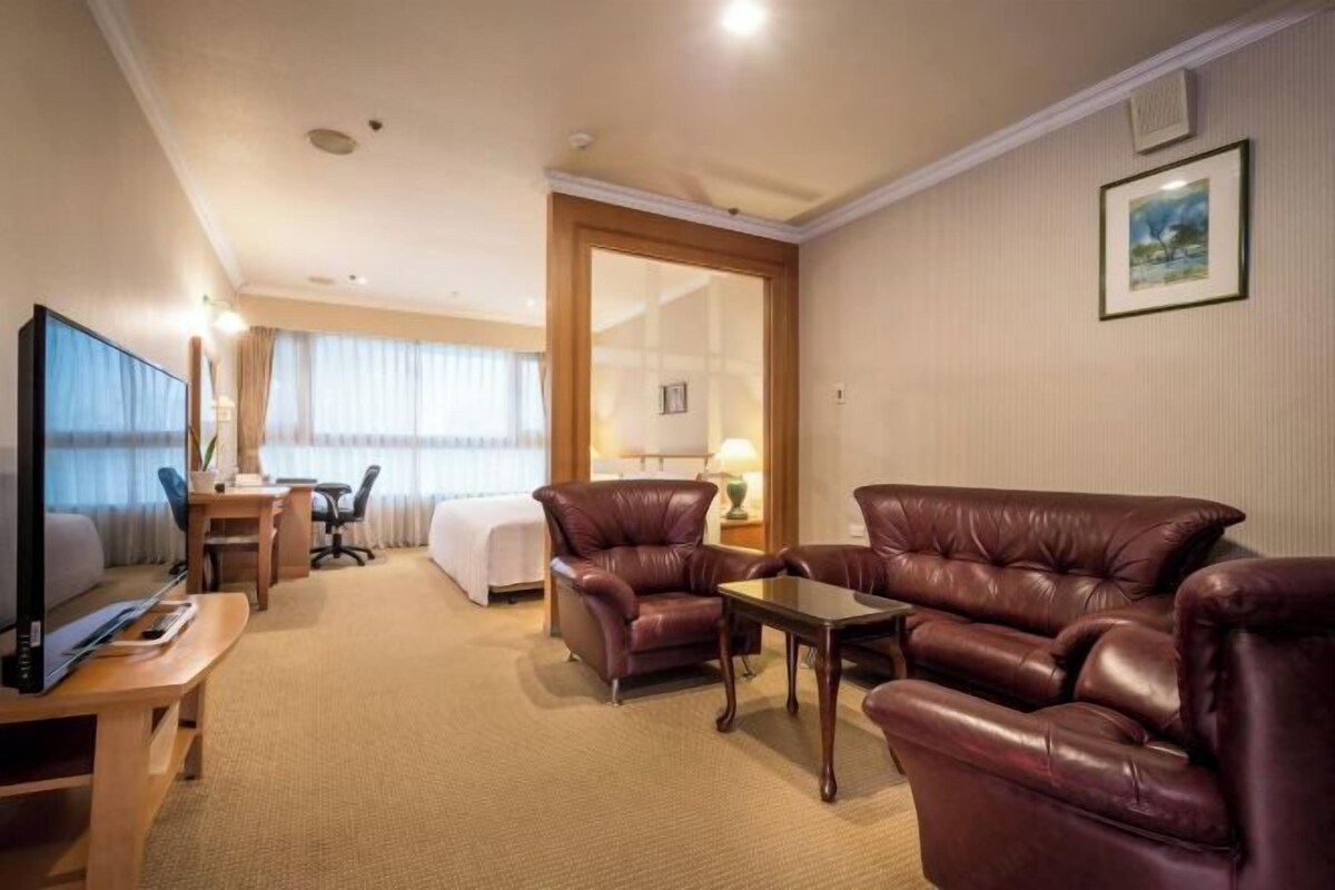 A hotel room with a bed, a desk, a TV, two leather armchairs, and a small table.