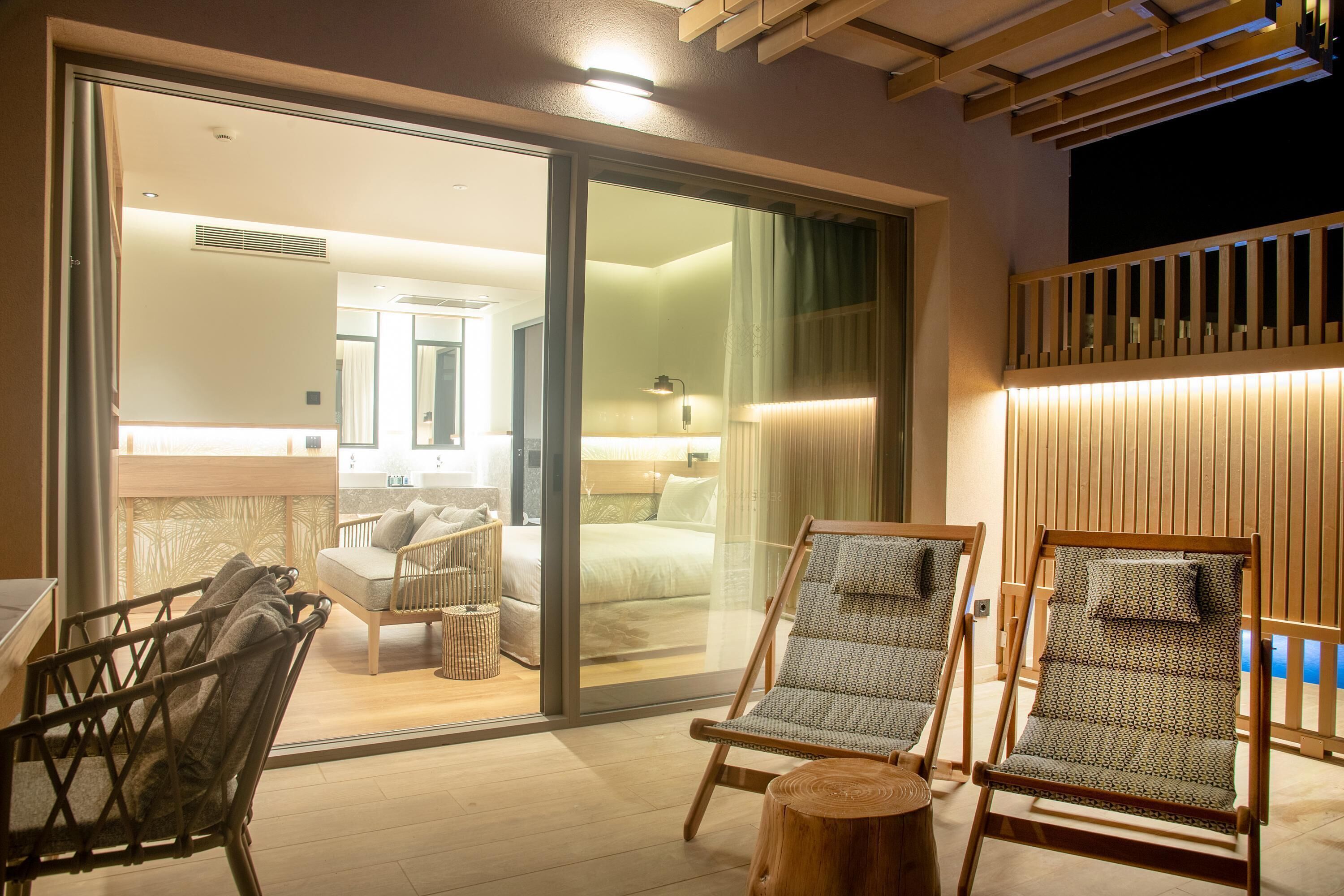 Una habitación de hotel moderna con un amplio ventanal corredero que da a un balcón, techo de madera y una cama con mesita de noche.