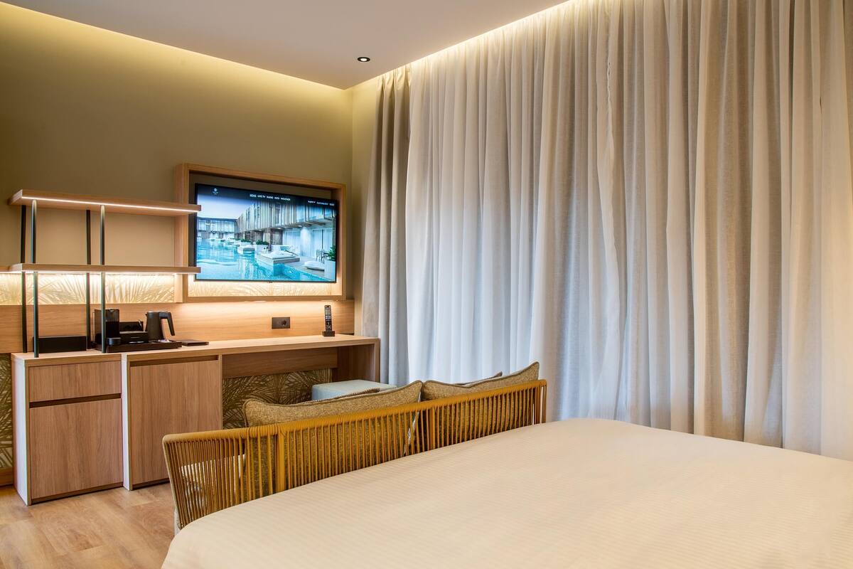 Une chambre d’hôtel moderne équipée d’un lit, d’un bureau avec un ordinateur, d’une télévision et d’une grande fenêtre avec des rideaux.