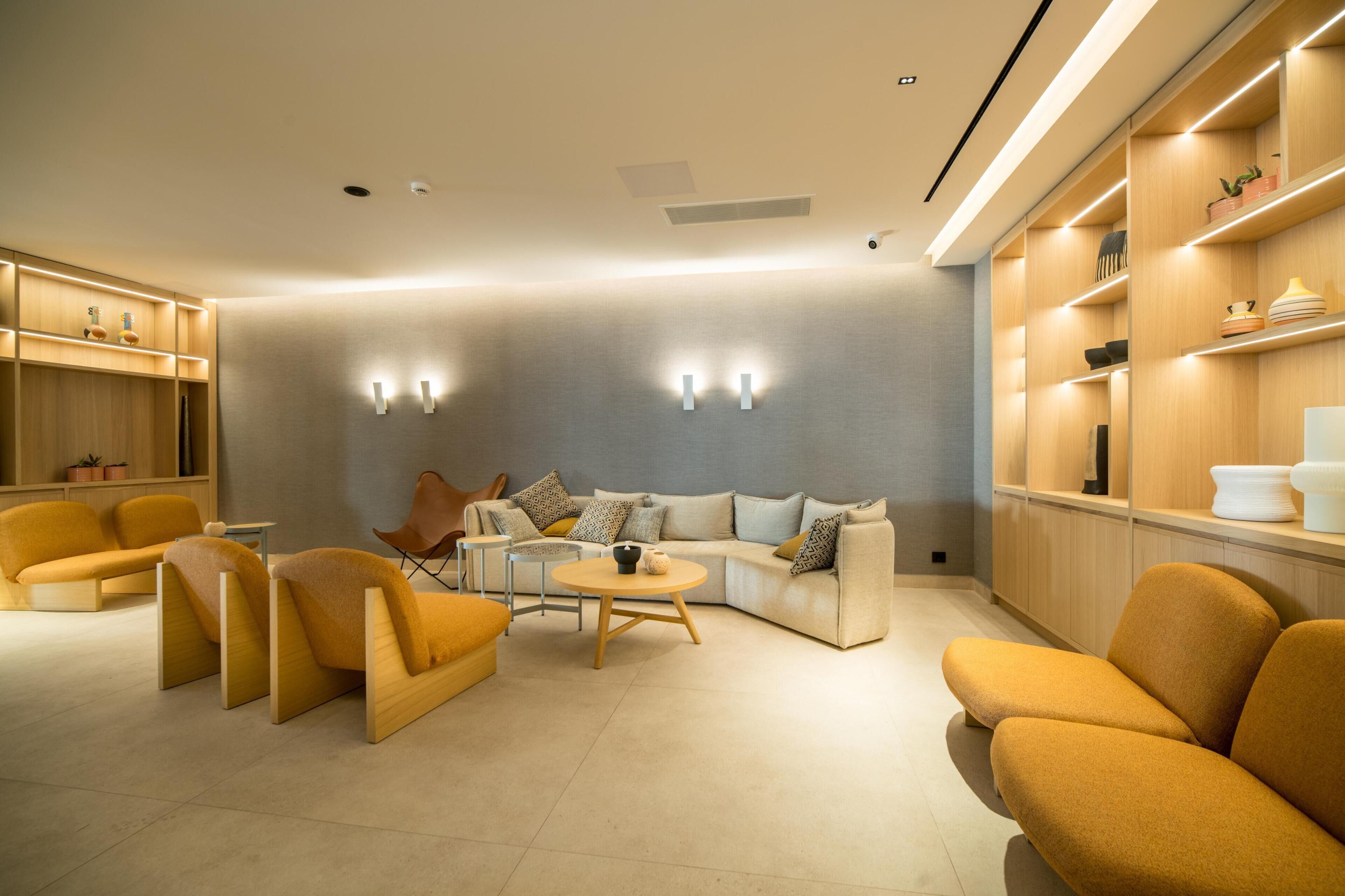 Ein modernes Wohnzimmer mit einer Couch, Sesseln und einem Couchtisch. Regale mit Dekoration und ein kleiner Tisch mit einer Lampe sind ebenfalls vorhanden.