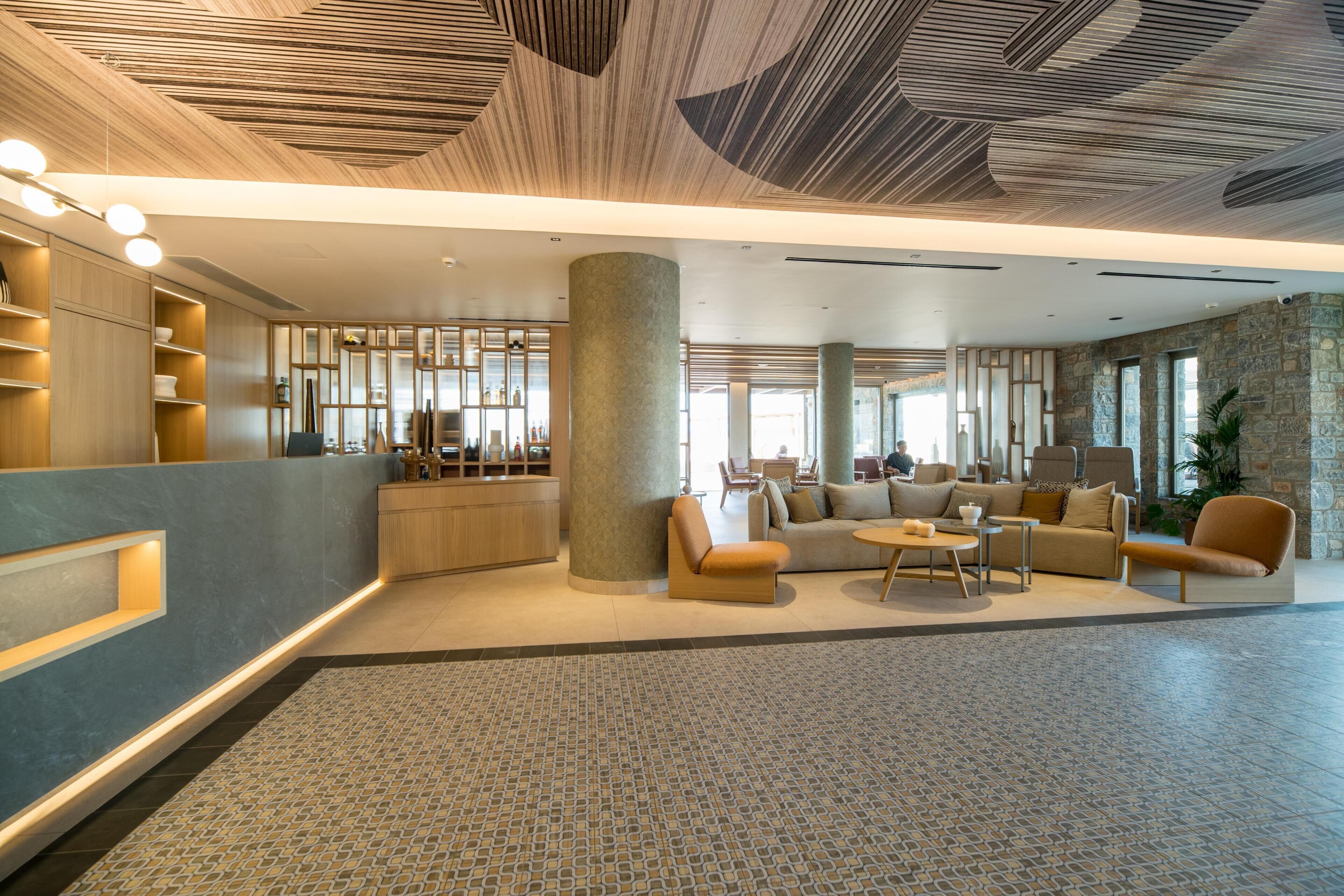 A spacious lobby with a modern design, featuring a central seating area with sofas and armchairs, a reception desk, and a patterned floor.