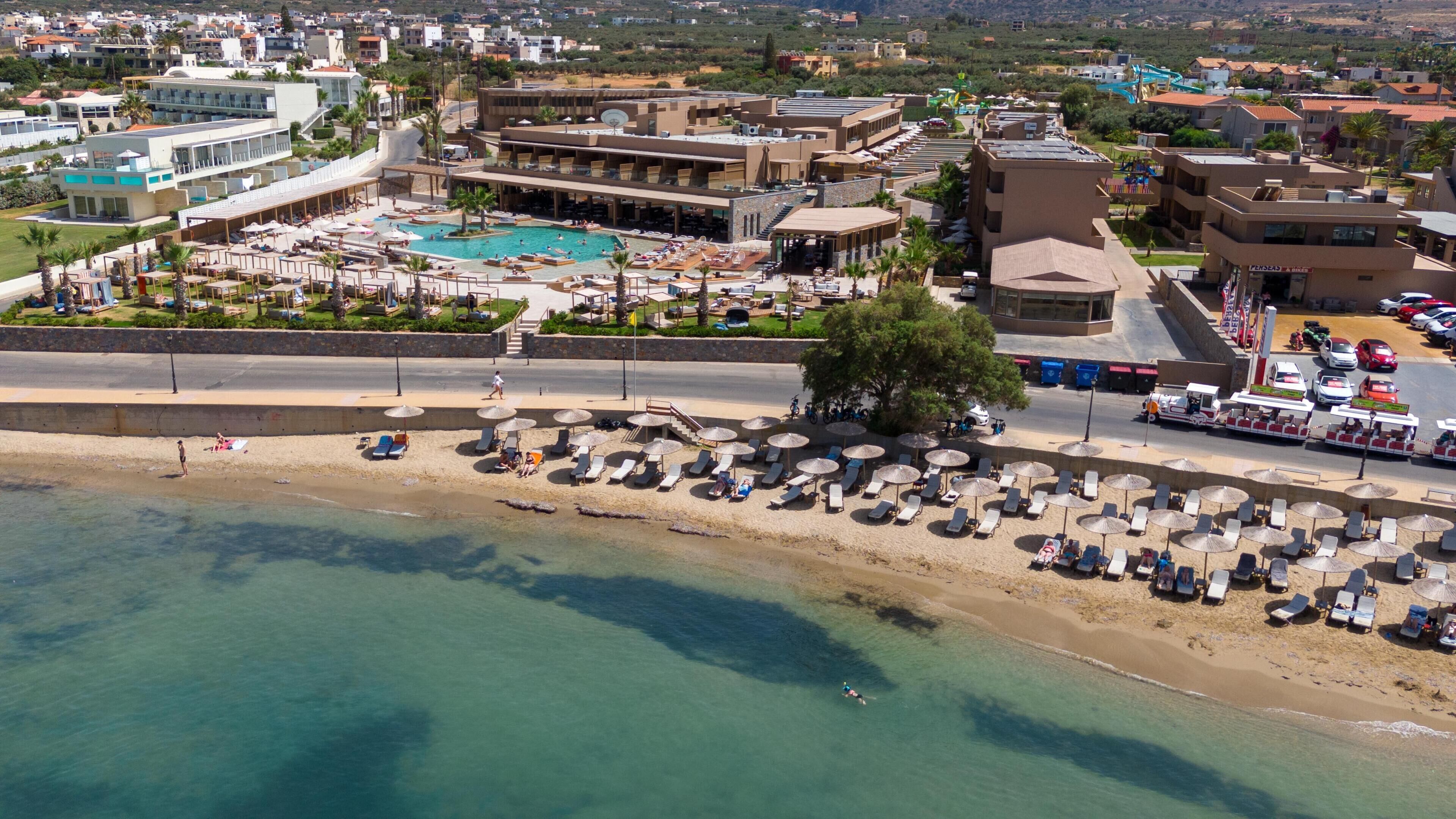 Vista aérea de un resort con una piscina grande, zona de playa con tumbonas y un estacionamiento.