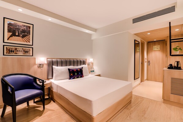 A modern hotel room with a large bed, a bedside table with a lamp, a chair, and framed pictures on the wall.