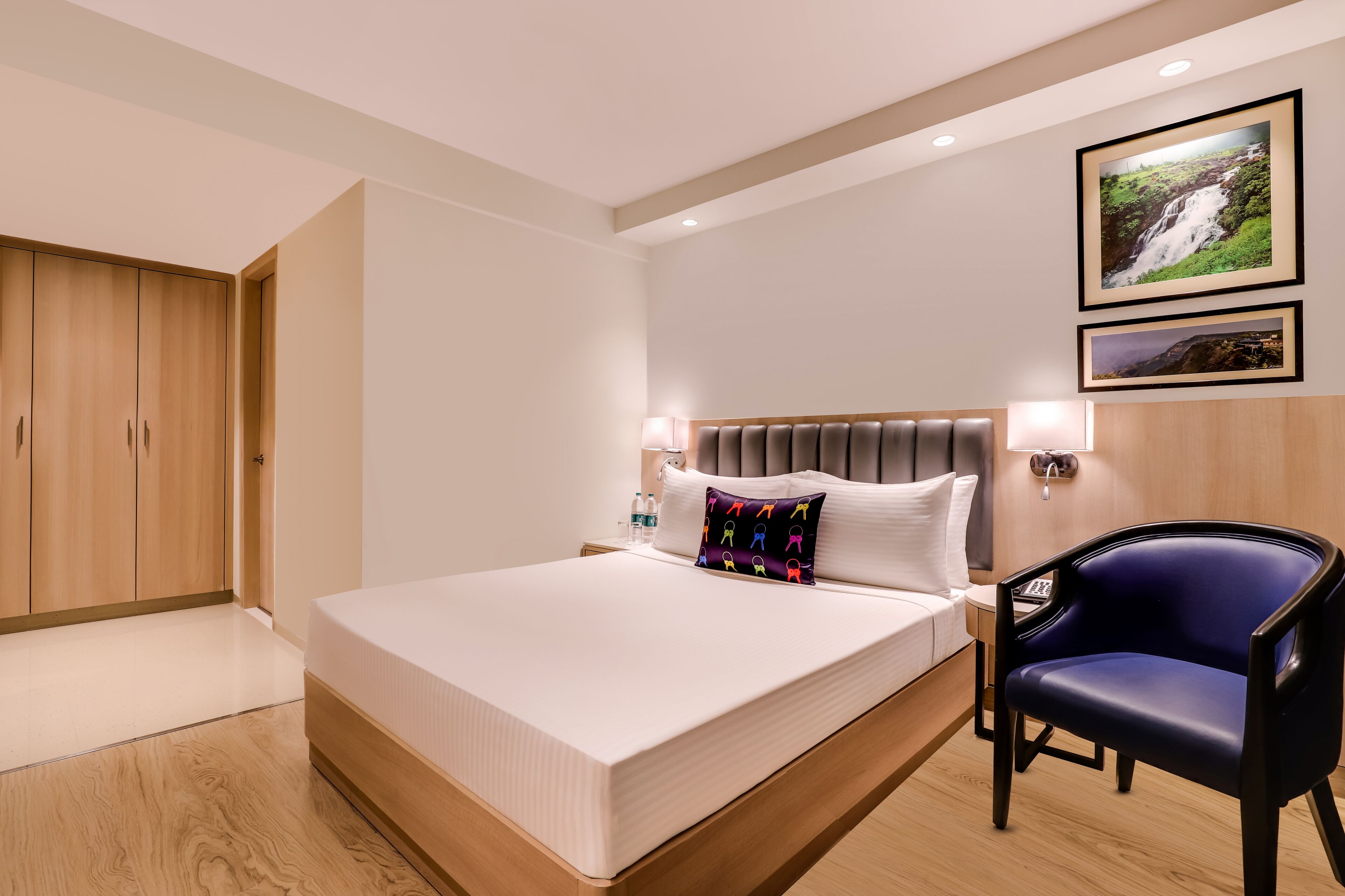 Quarto de hotel moderno com uma cama grande, um guarda-roupa de madeira, uma cadeira azul e duas molduras na parede.