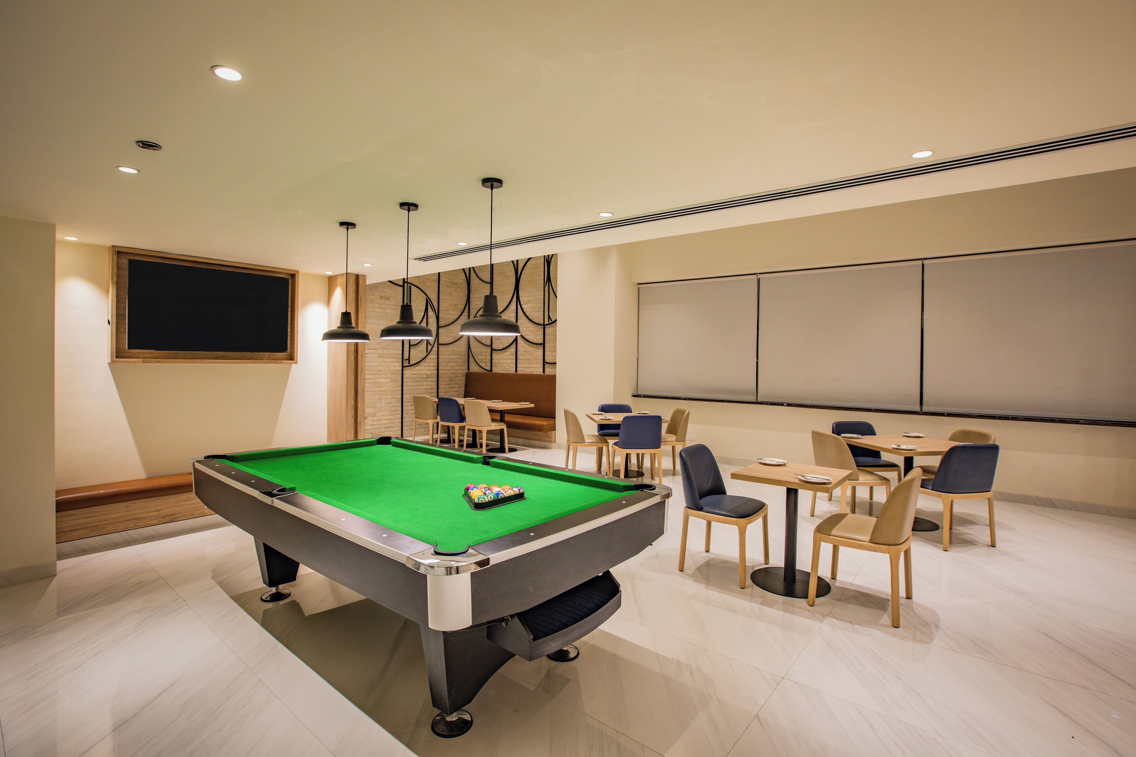 A pool table with a green felt surface, surrounded by chairs and tables in a well-lit room with recessed ceiling lights.