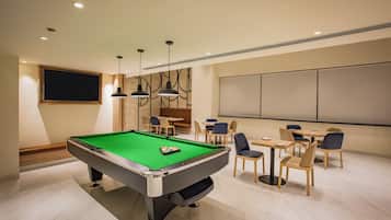 A pool table with a green felt surface, surrounded by chairs and tables in a well-lit room with recessed ceiling lights.