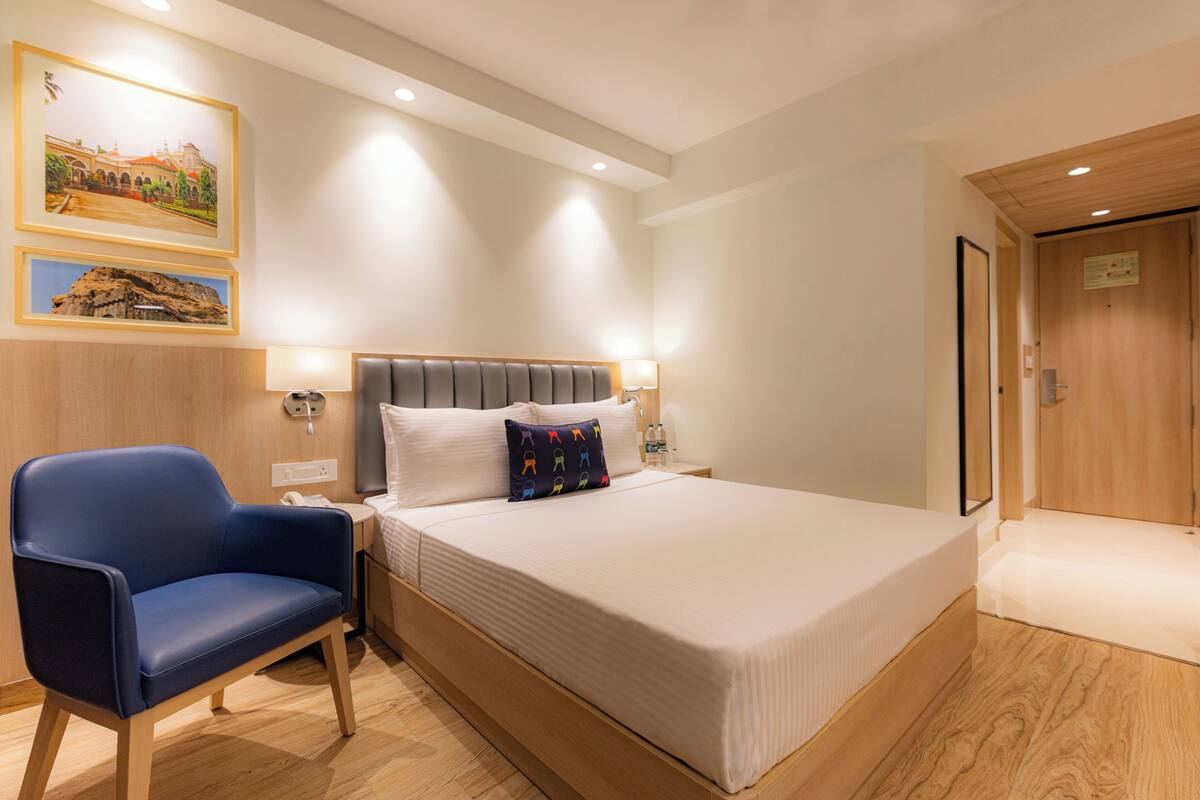 Una habitación de hotel con una cama, una silla azul, una mesita de noche, lámparas fijadas a la pared y cuadros enmarcados.
