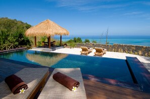 A poolside area with lounge chairs, a thatched umbrella, and a view of the ocean. - Lakaz Chamarel Exclusive Lodge (Chamarel)