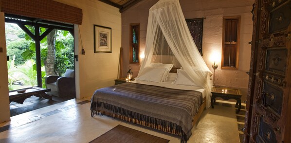 Un dormitorio con cama con dosel, una mesita de noche con lámpara, una ventana con vistas a la vegetación y una puerta con detalles de metal ornamental.