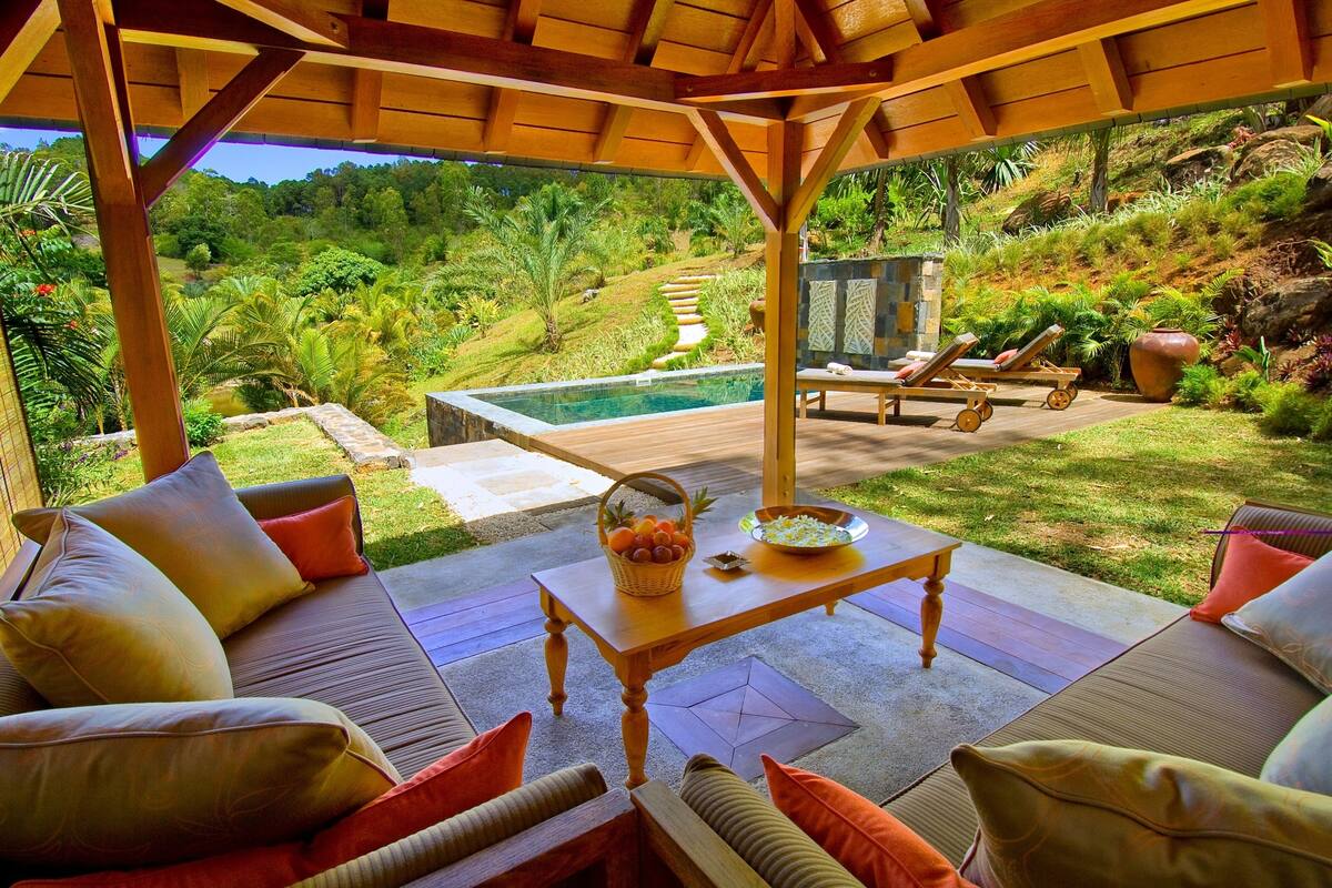 Uma área de estar externa com piscina, cercada por vegetação exuberante e uma pérgula de madeira.