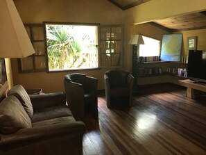 A cozy living room with a leather sofa, a wooden coffee table, a bookshelf, a television, and a large window with a view of greenery. - Lakaz Chamarel Exclusive Lodge (Chamarel)