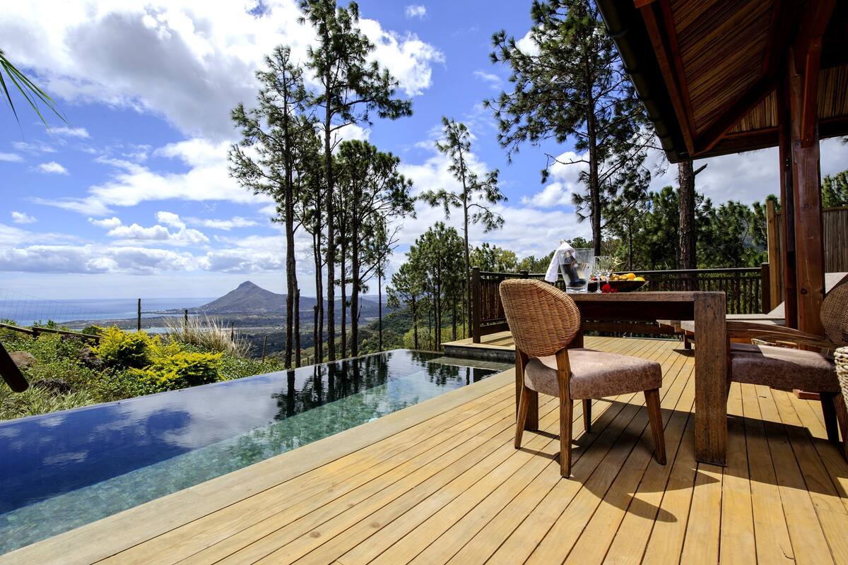 Uma piscina infinita com deck de madeira, cadeiras de vime e uma mesa, com uma pessoa ao fundo.