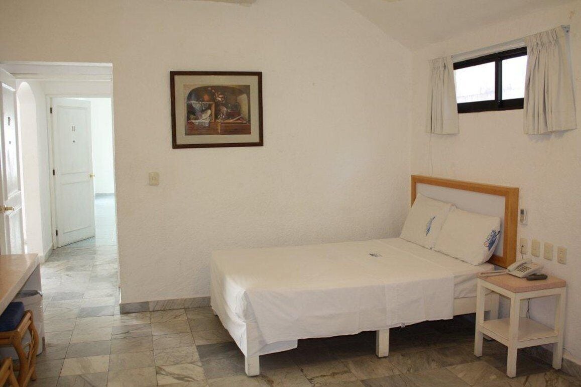 Una habitación pequeña y limpia con una cama individual, una mesita de noche con teléfono, un cuadro en la pared y una ventana con cortinas.