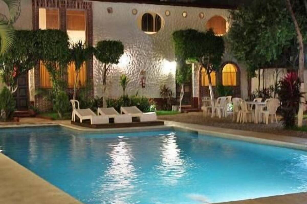 Piscina con sillas de descanso y mesas, rodeada de follaje y elementos arquitectónicos.