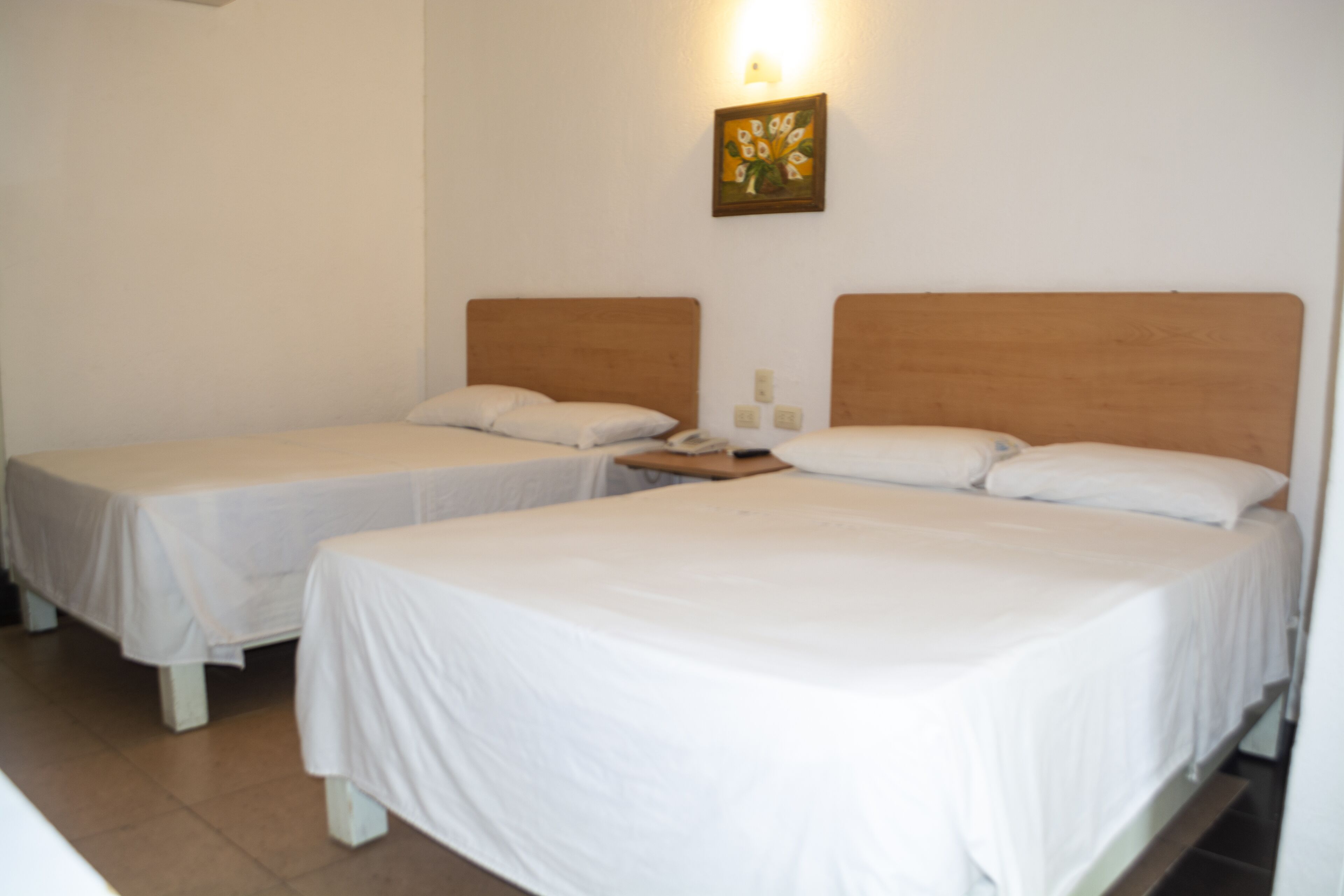 A hotel room with two single beds, a wooden headboard, a small bedside table with a lamp, and a wall-mounted painting.