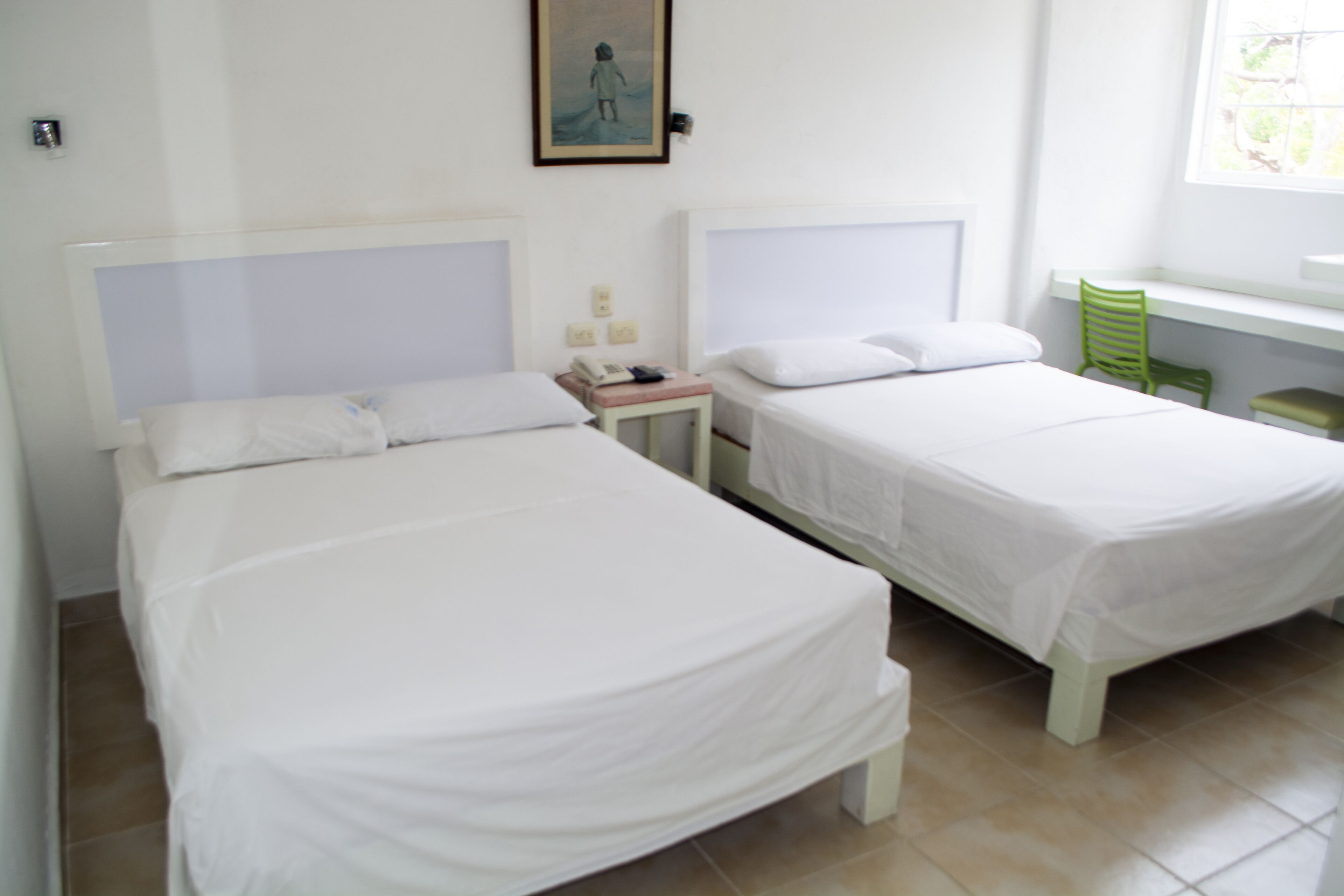 A hotel room with two single beds, a small bedside table with a phone, a green chair, and a window with a view of trees.