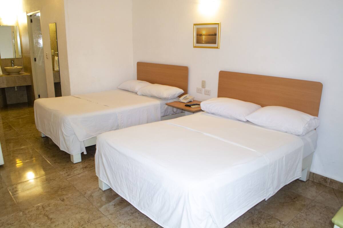 Habitación de hotel con dos camas individuales, cabecera de madera, mesita de noche con teléfono y baño con lavamanos y espejo.