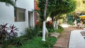 Un sendero en un jardín que lleva a un edificio blanco con parte de ladrillo, rodeado de abundante vegetación y un patio con mobiliario exterior.