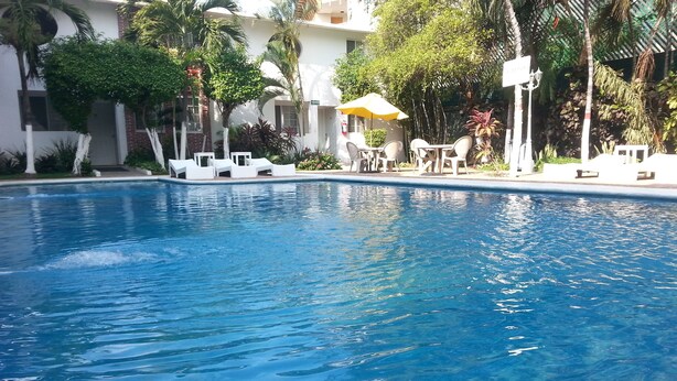 Piscina con sillones de descanso y sombrillas, rodeada de palmeras y un edificio de fachada blanca.