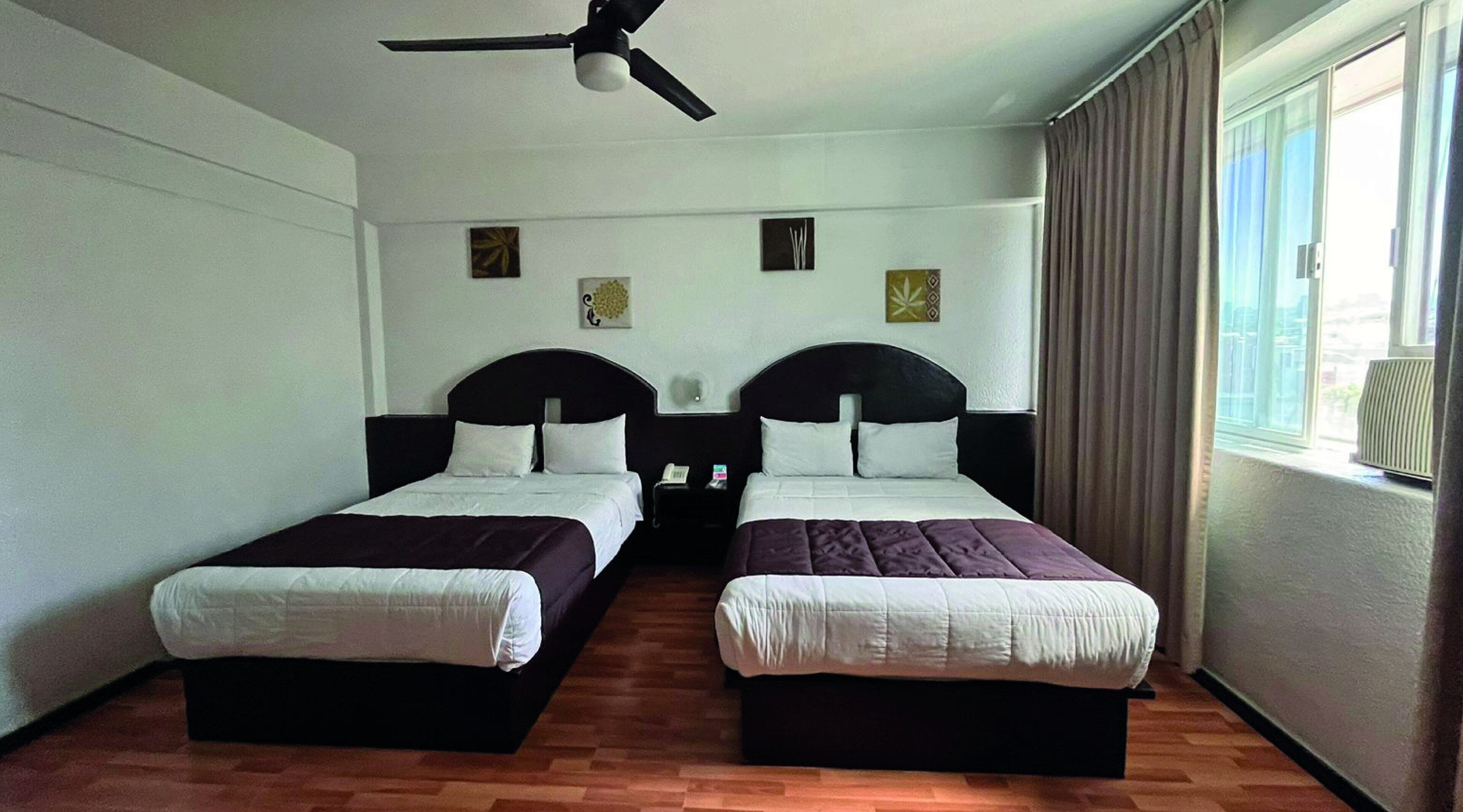 A hotel room with two single beds, a ceiling fan, a bedside table with a phone, and a window with curtains.