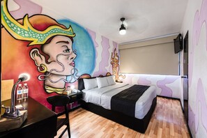 A bedroom with a bed, a desk with a lamp, a chair, and a wall mural. - HI Hotel Impala (Querétaro)