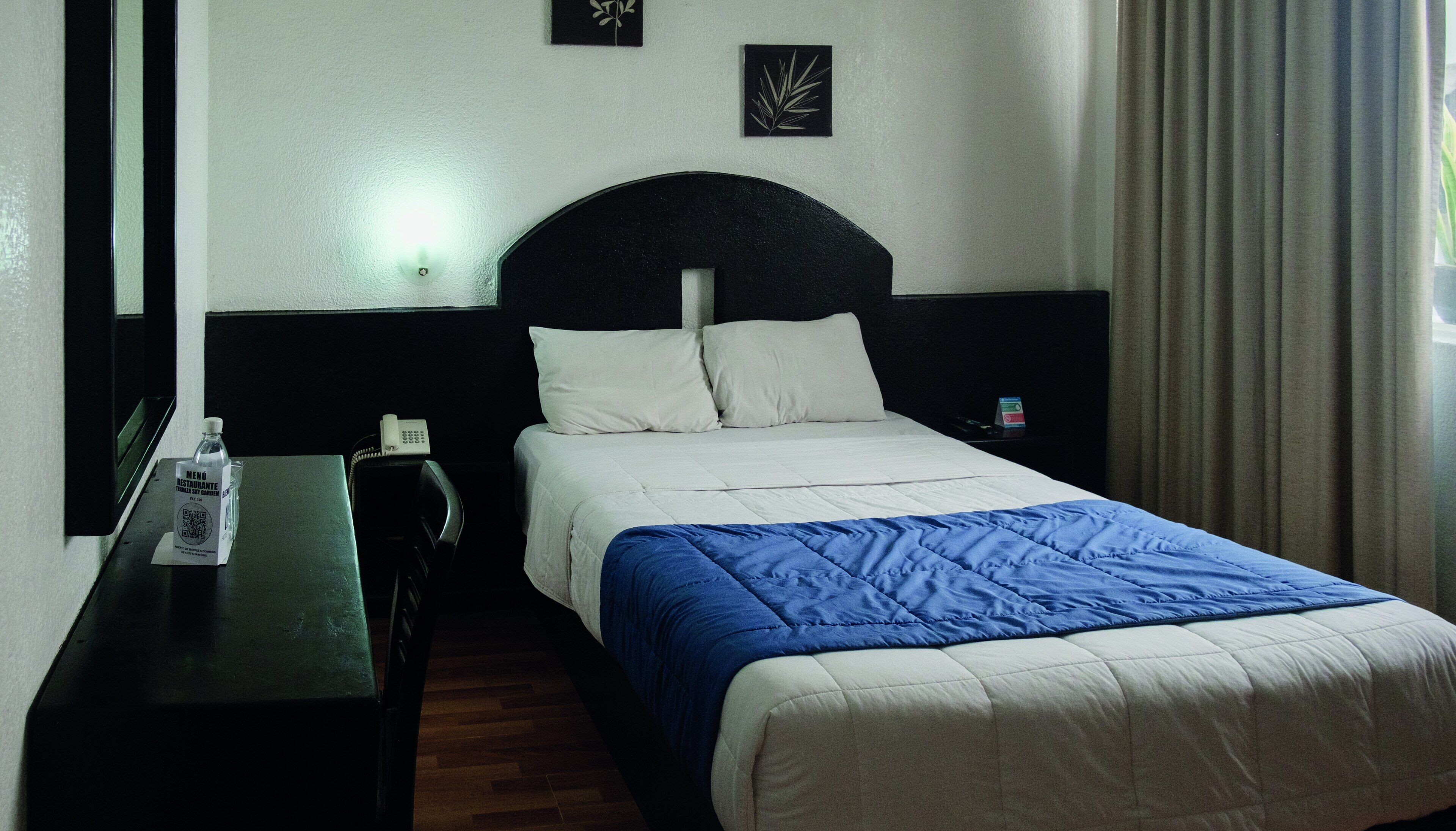 A hotel room with a bed, a nightstand, a telephone, and a small table with a bottle and a menu.