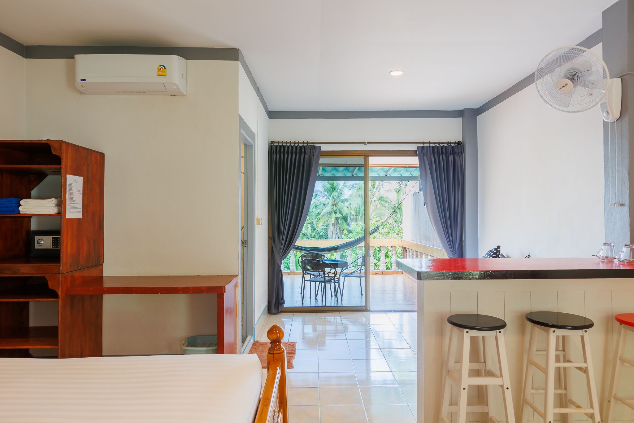 A hotel room with a bed, a wooden desk, a kitchenette, and a balcony with a view of palm trees.
