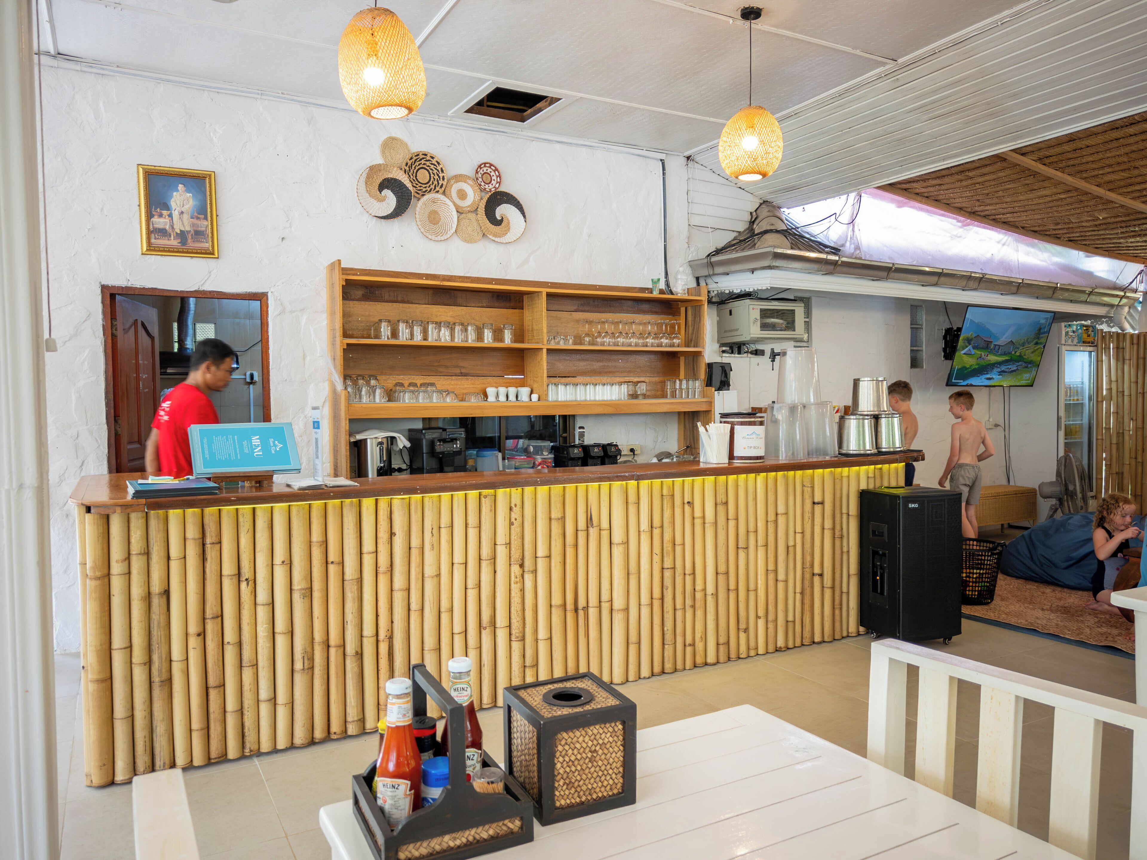 Een restaurantinterieur met een houten toonbank, planken met glazen en kopjes, een televisie en een bamboewand.