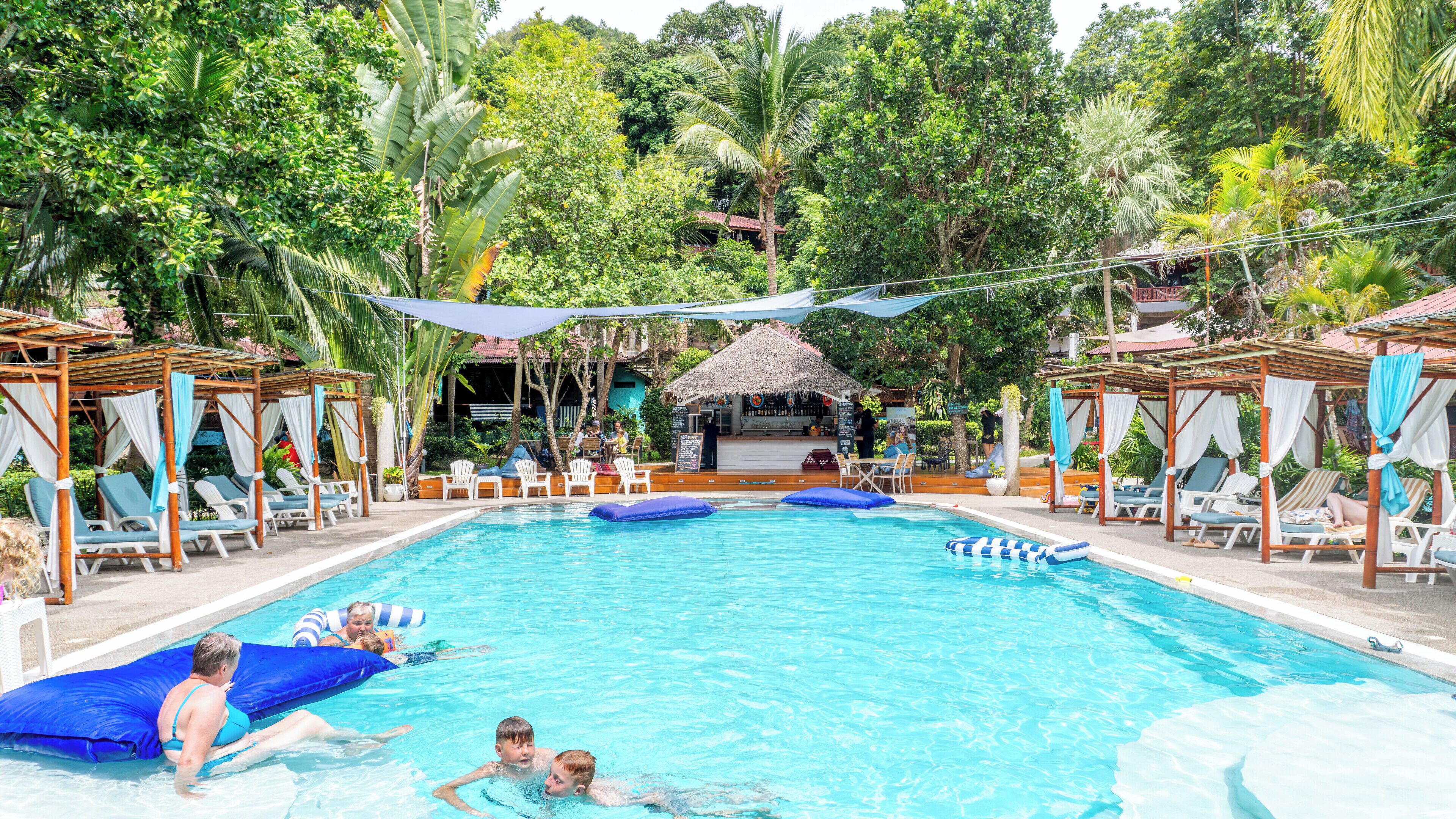 Piscina con sillas de descanso y sombrillas, rodeada de exuberante vegetación y una estructura de techo de paja.