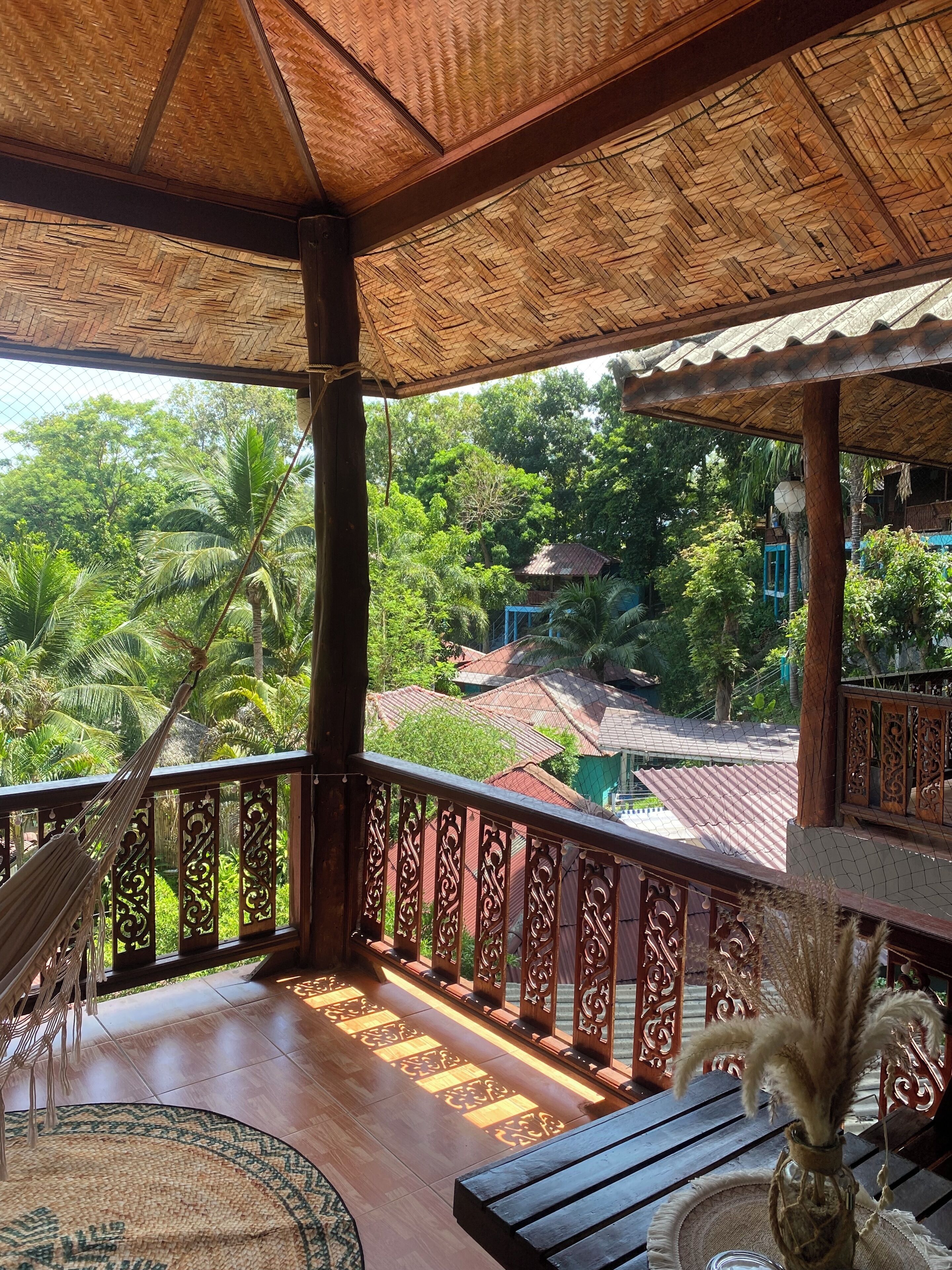 A wooden balcony with a thatched roof, a hammock, and a view of lush greenery and buildings in the distance.