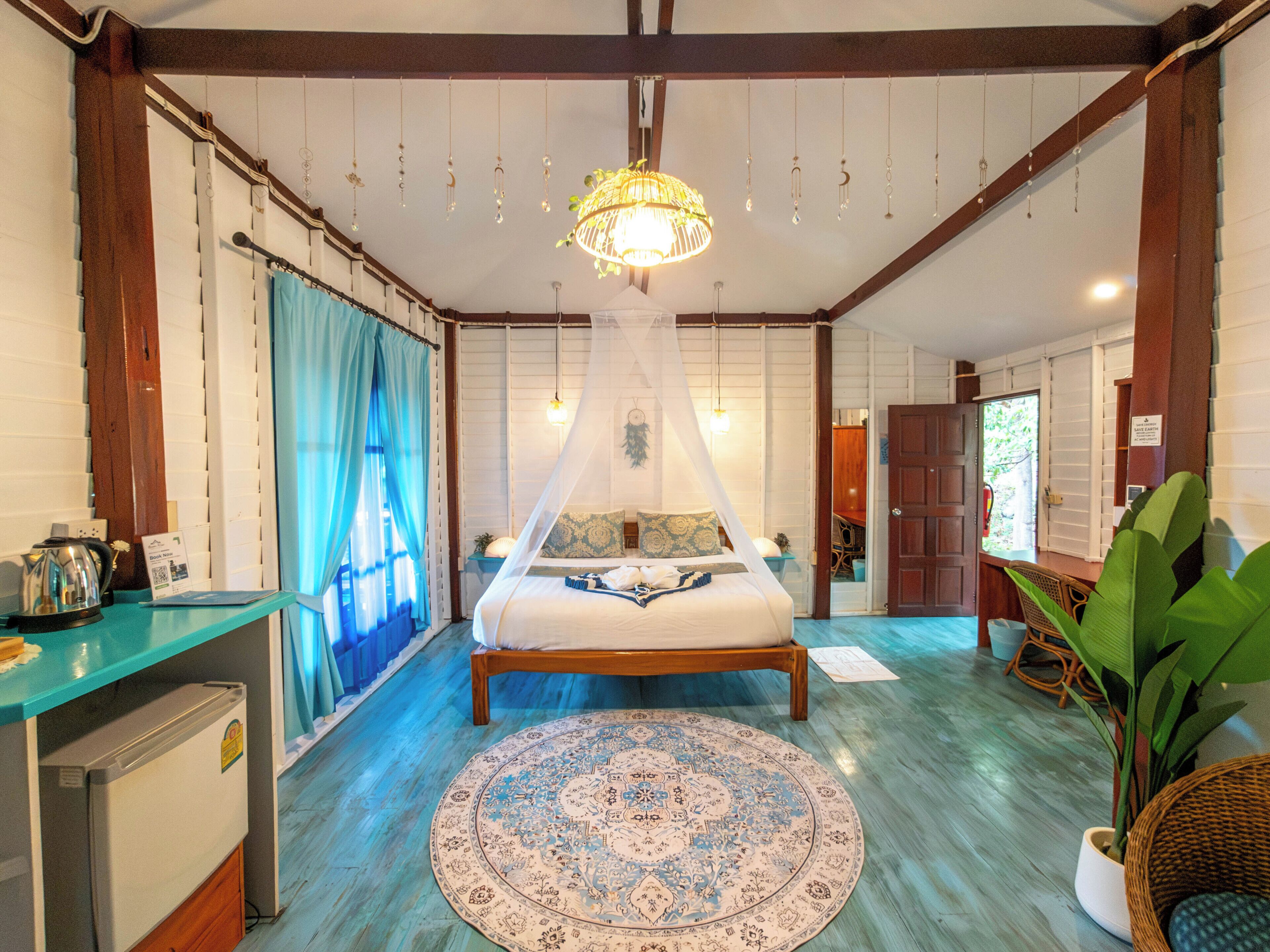 A room with a bed, a round patterned rug, a small table with a kettle, and a large plant.