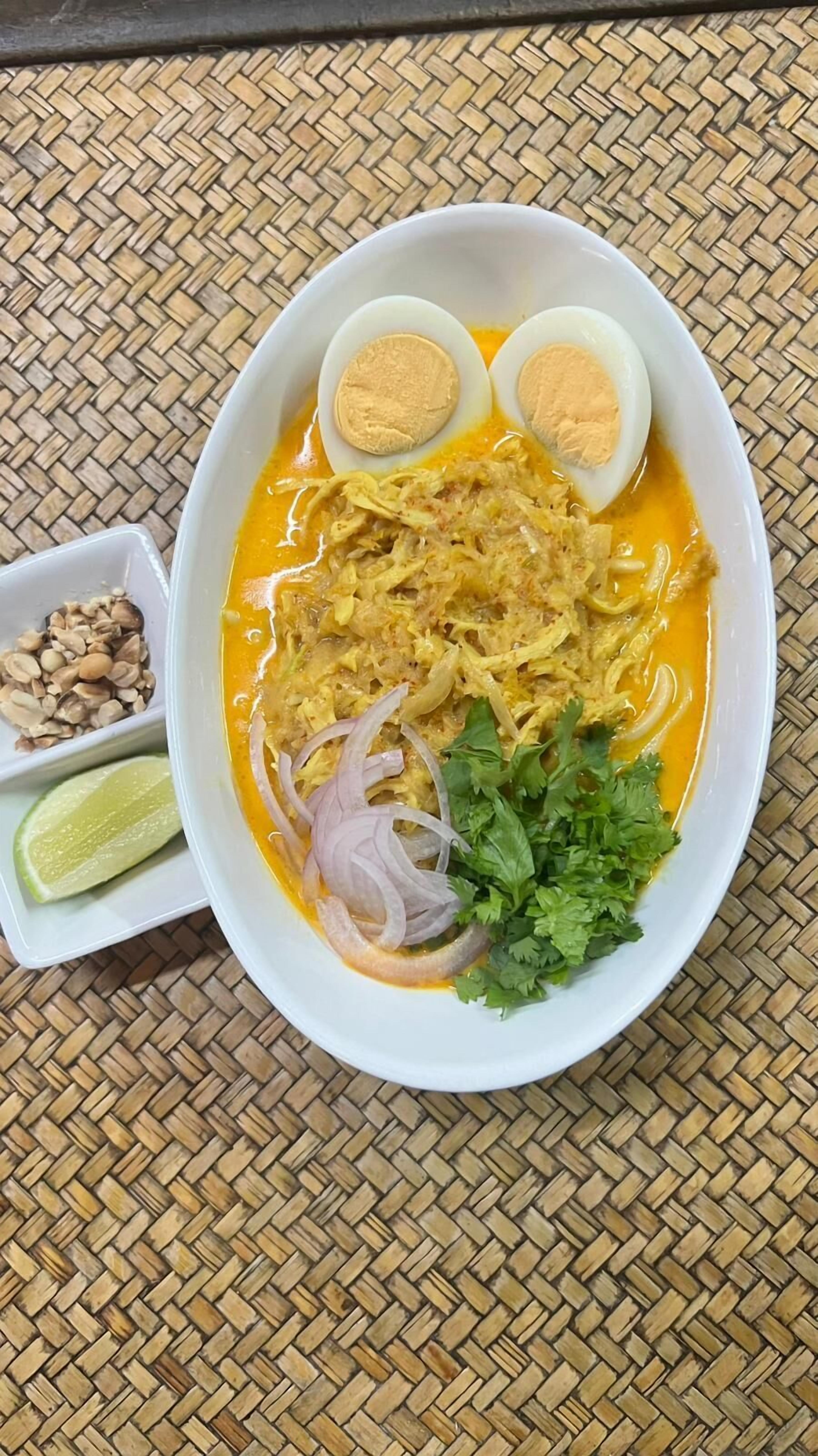 A bowl of noodle soup with boiled eggs, shredded chicken, onions, cilantro, and a lime wedge on a woven mat.