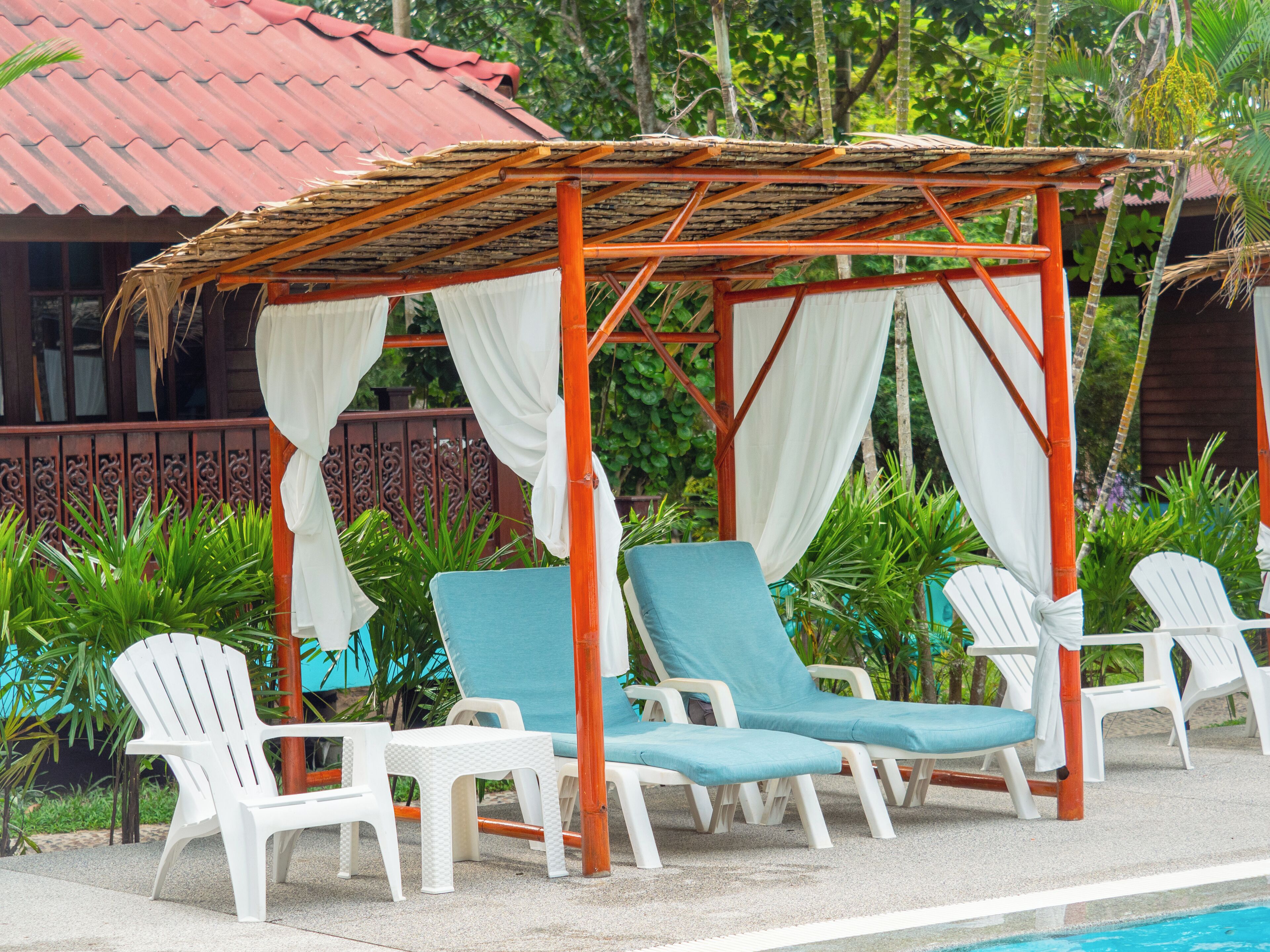 A covered outdoor seating area with white plastic chairs and a pool in the background.