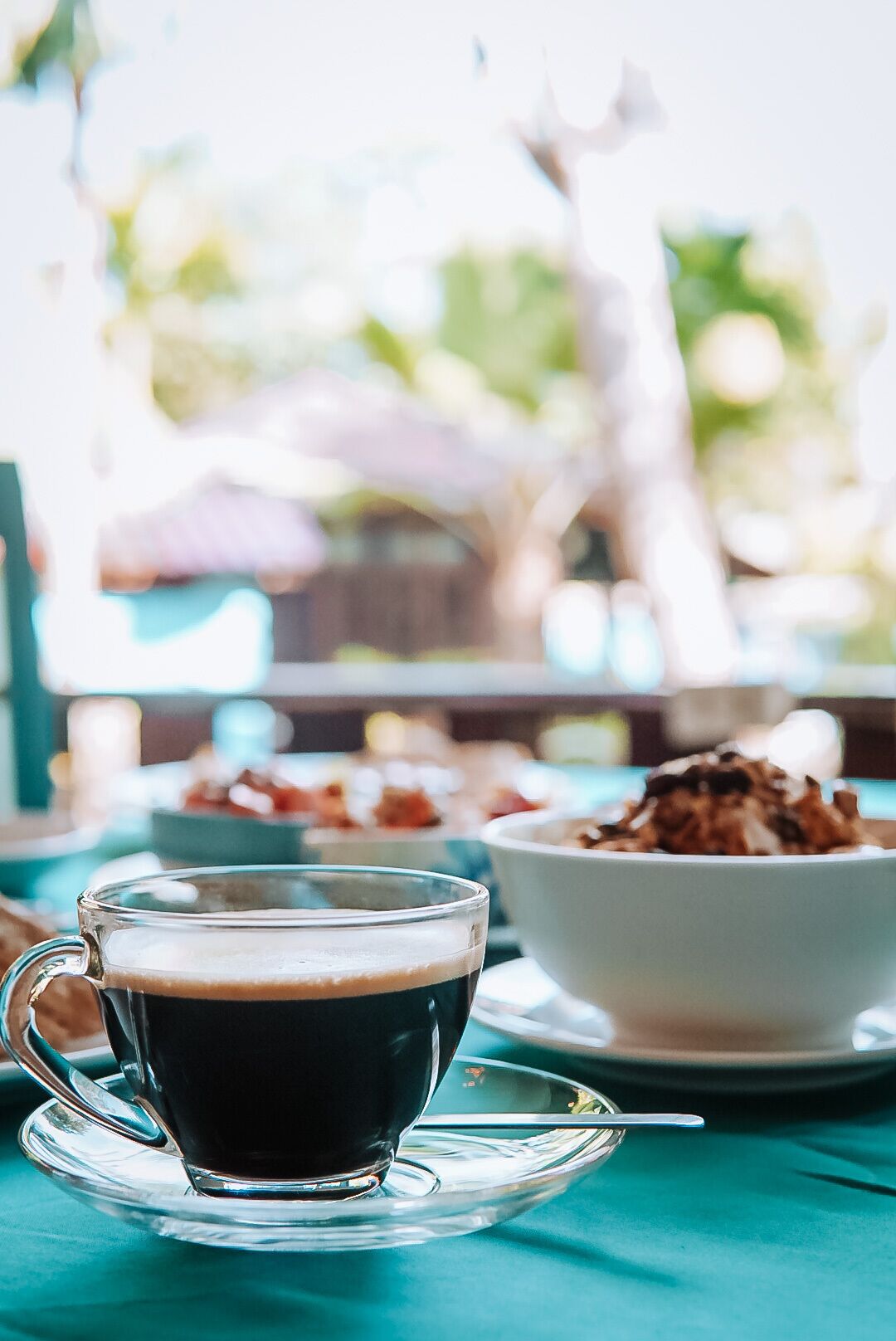 Une tasse de café en verre, posée sur une soucoupe avec une cuillère, sur une table recouverte d’une nappe de couleur turquoise.