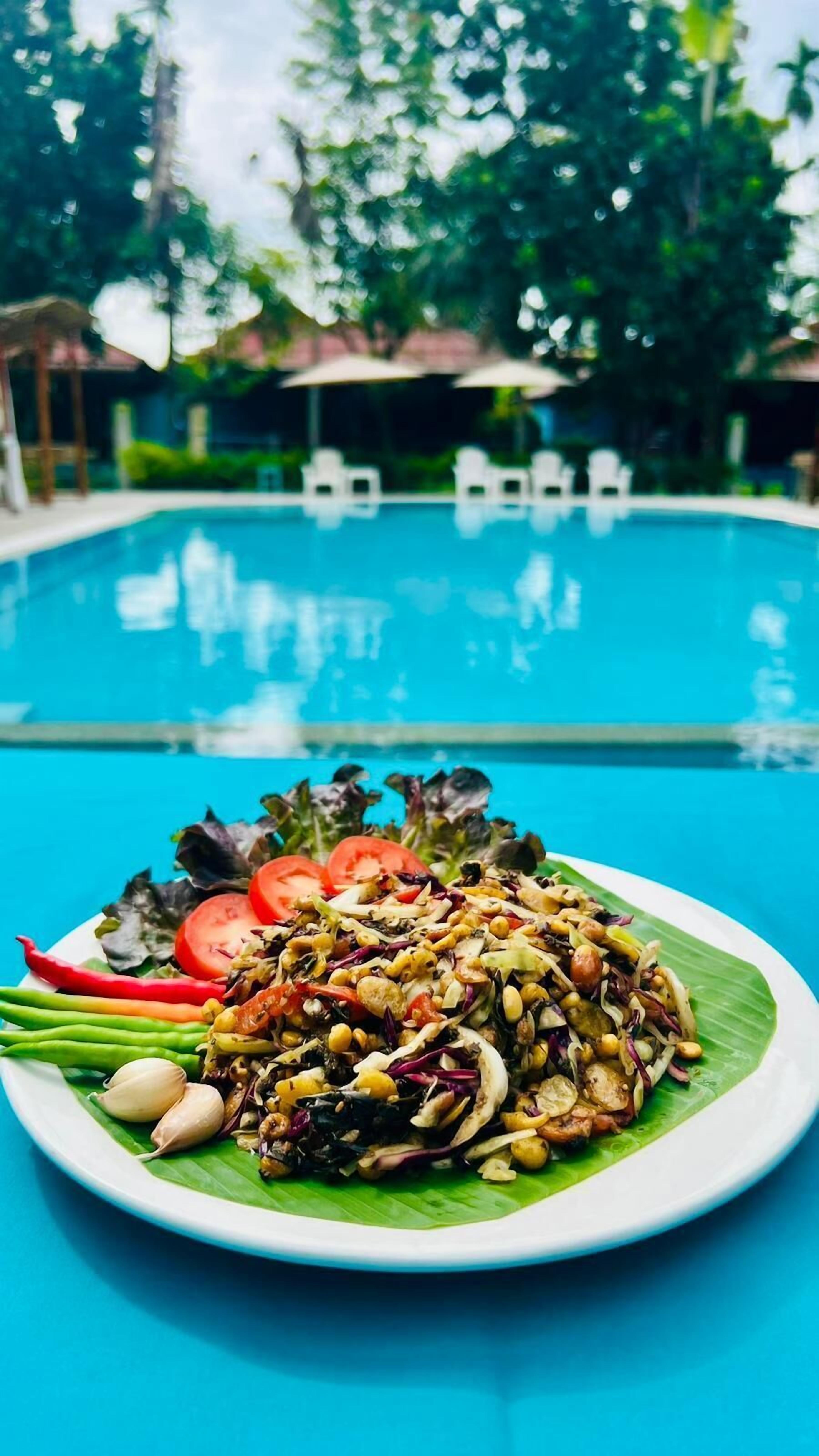 A plate of salad with various vegetables and nuts on a white plate, placed in front of a swimming pool with lounge chairs in the background.