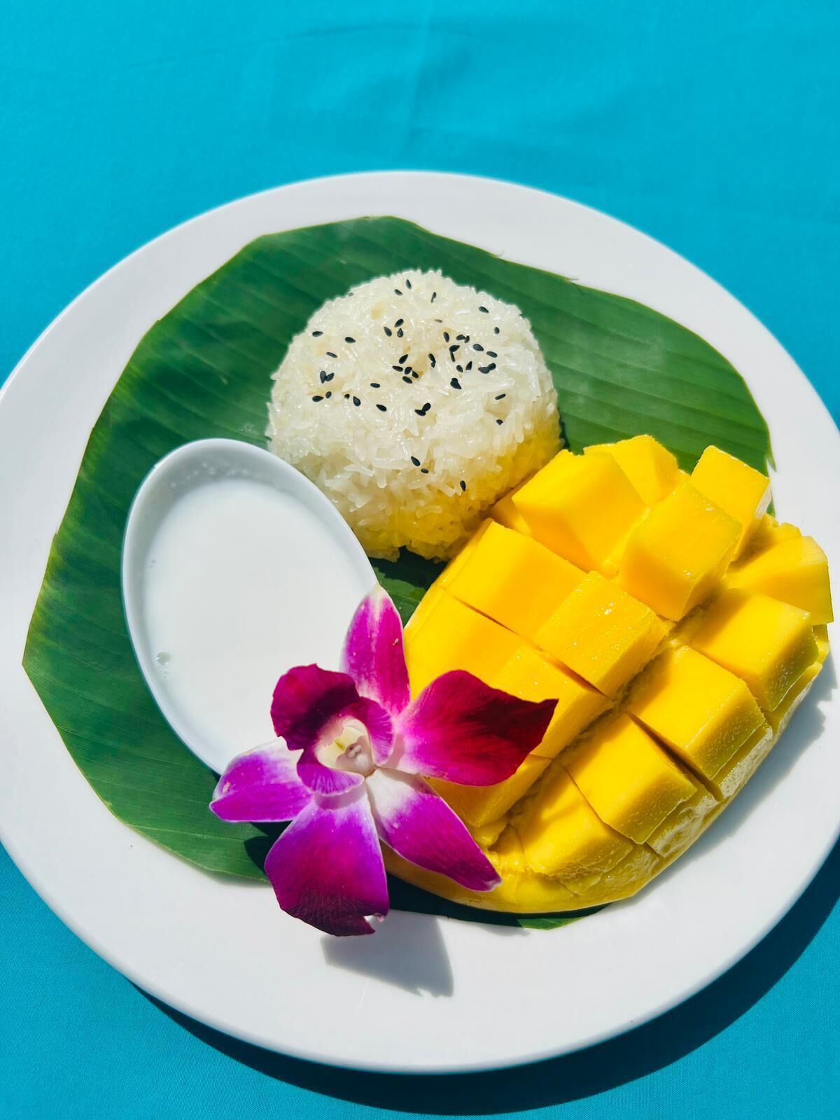 Un plato con arroz blanco cubierto de semillas de sésamo negro, cubos de mango amarillo y un pequeño recipiente blanco, todo servido sobre una hoja verde.