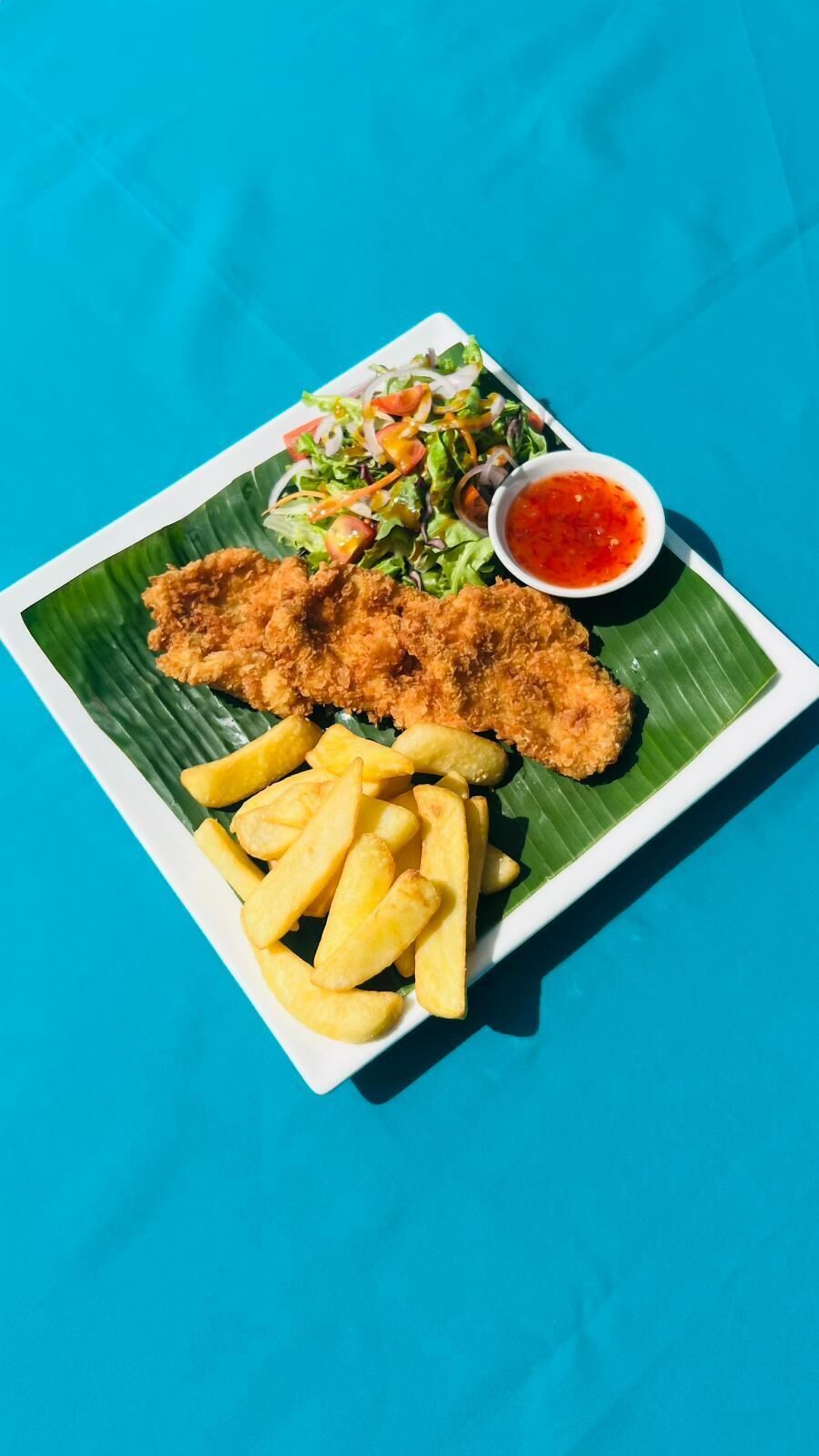 A plate of fried fish, French fries, a salad, and a side of red sauce on a white plate with a green leaf underneath.