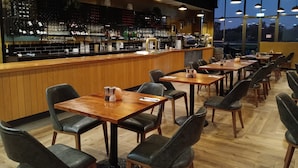 A restaurant with wooden tables and black leather chairs, a bar area with shelves of bottles and glasses, and large windows offering a view of the outside. - Quality Hotel Point Cook (Point Cook)