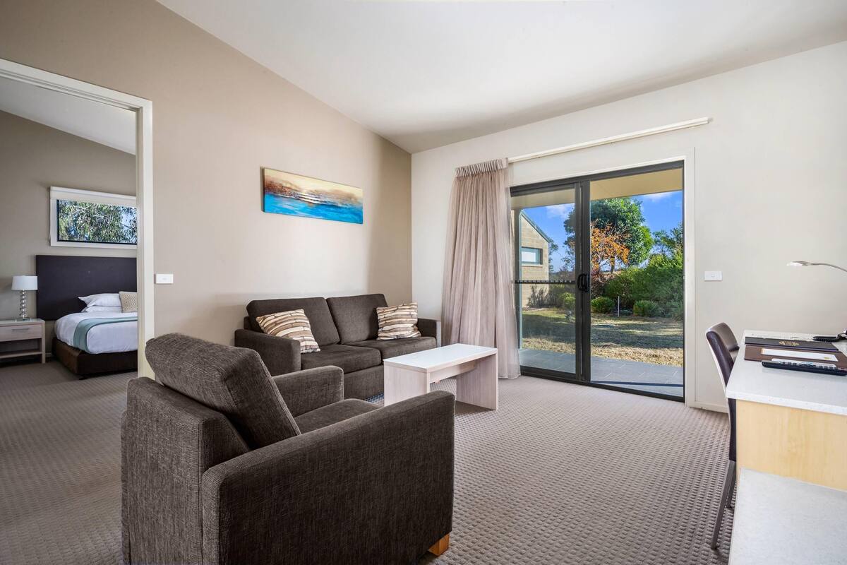 A modern hotel room with a sofa, a small table, a desk, a bed, and a large sliding glass door leading to a balcony.