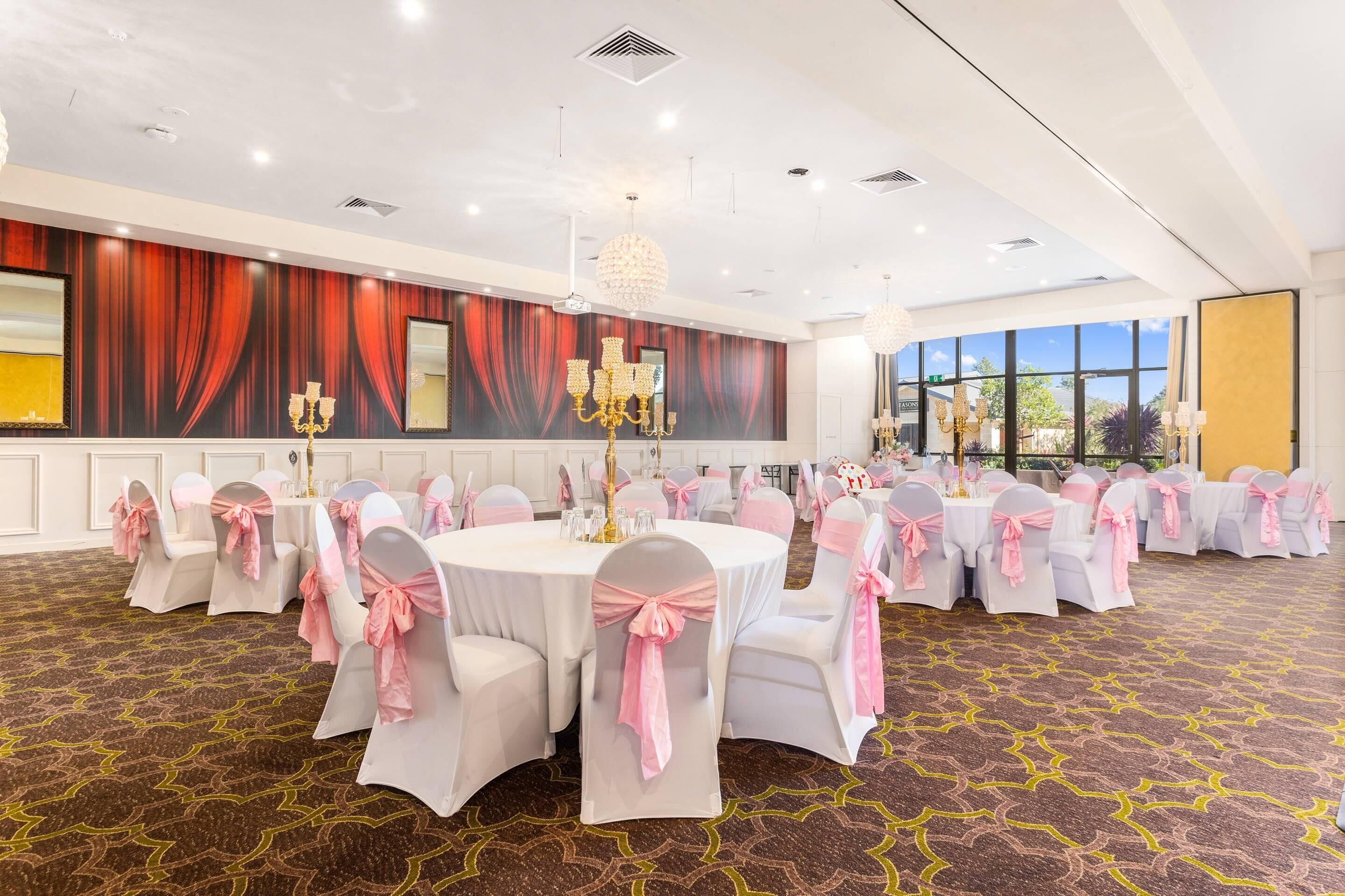 Een feestzaal met ronde tafels, stoelen met roze linten, kroonluchters en een groot raam met uitzicht.
