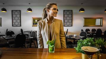 A woman seated at a wooden bar in a restaurant with multiple tables and chairs, framed artwork on the walls, and pendant lighting.