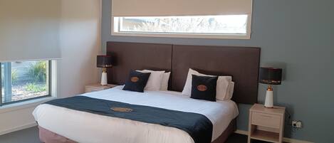 Una cama bien hecha con cabecera, dos mesitas de noche con lámparas, una ventana con persianas y vistas al exterior.
