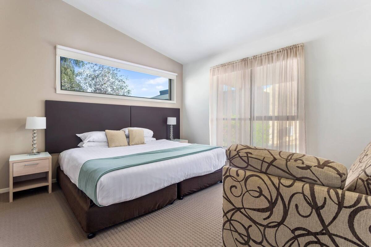 A hotel room with a bed, a nightstand, a chair, a window with curtains, and a framed picture on the wall.