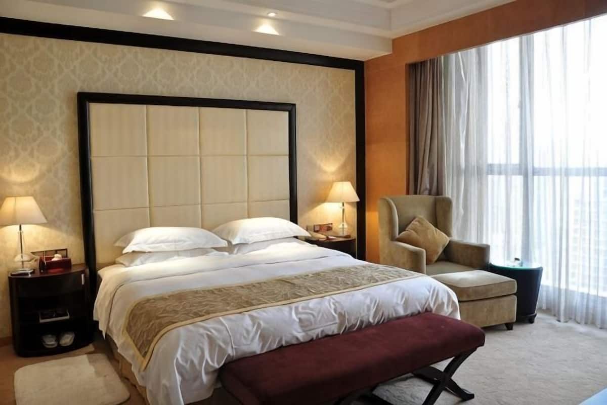 Quarto de hotel com uma cama grande, duas mesas de cabeceira com abajures, uma cadeira, um banco e uma janela ampla com cortinas.